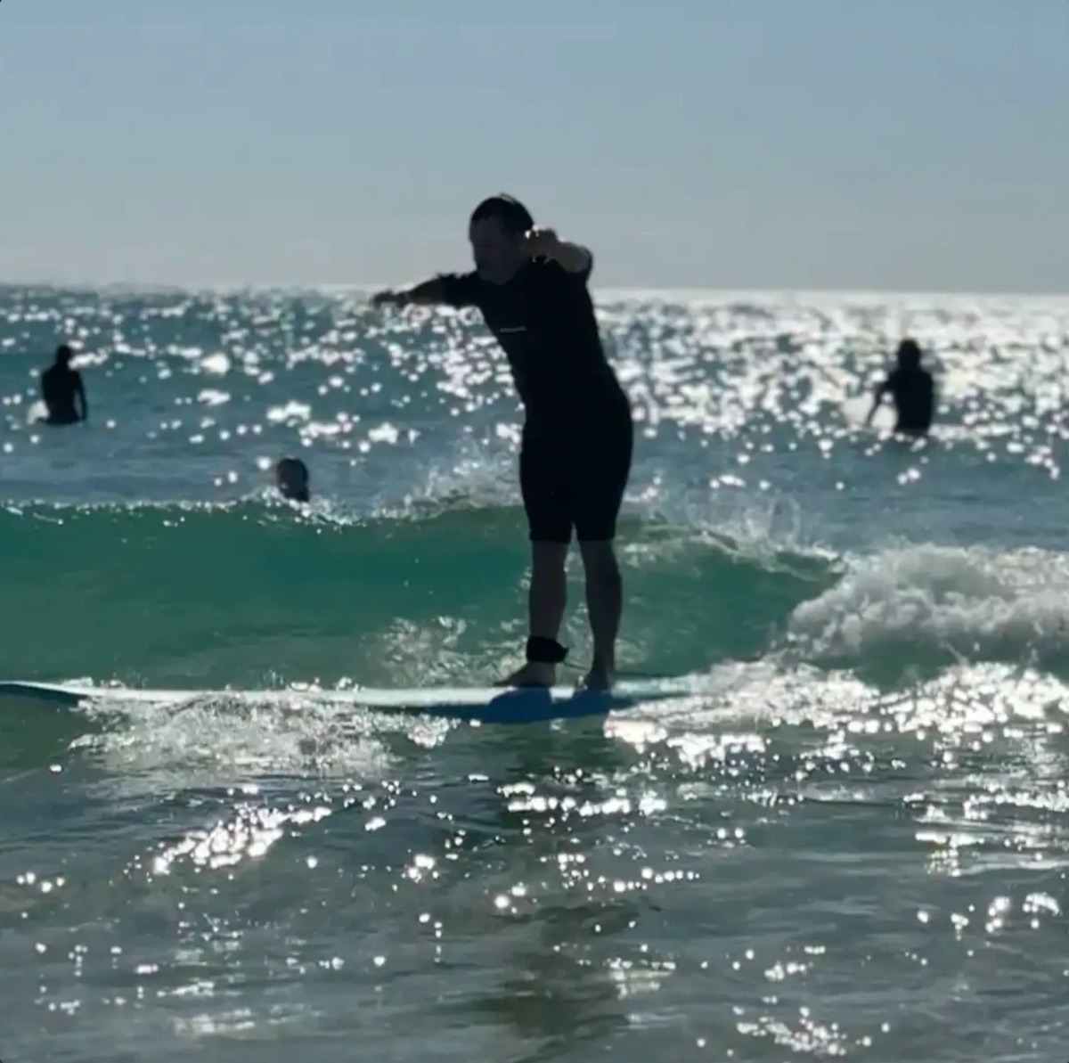 A surfer silhouetted against the glare, standing up on a board as a small wave rolls in